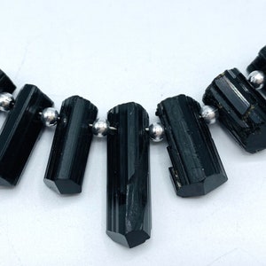 May include: A necklace featuring seven black tourmaline gemstone beads, each with a cylindrical shape and faceted ends. The beads are strung on a thin, silver-colored wire, interspersed with small, round, silver-colored beads.
