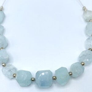May include: A strand of aquamarine gemstone beads. The beads are a pale blue color, with irregular, faceted shapes. Each bead is separated by a small, gold-colored spacer. The beads are strung on a thin, clear cord.