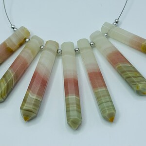 May include: A necklace featuring seven elongated, faceted gemstone pendants. The pendants display bands of white, pink, green, and yellow hues. Each is attached to a silver-colored bead and strung on a thin, silver-colored wire.