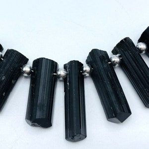 May include: A necklace featuring seven black tourmaline gemstone beads, each approximately 1 inch long, strung on a silver-colored wire. Silver beads are interspersed between the tourmaline pieces. The background is white.