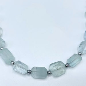 Pode incluir: Um colar com contas facetadas de pedra preciosa água-marinha azul clara. As contas são enfiadas com pequenas contas de metal prateado. As pedras preciosas têm uma qualidade translúcida e são de forma irregular.