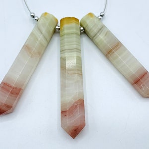 Pode incluir: Três varinhas de cristal de ônix polidas. As varinhas são de cor creme claro com faixas vermelhas e marrons. Cada varinha tem uma extremidade facetada e pontiaguda. As varinhas são enfiadas em um fio de prata fino.