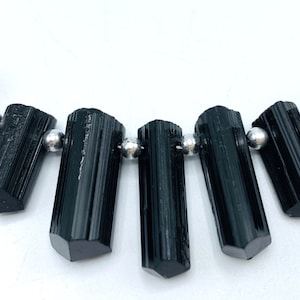 May include: A strand of black tourmaline gemstone beads. The cylindrical, polished stones are strung with small silver-colored beads. The tourmaline crystals have a glossy finish and are arranged in a gentle curve against a white background.