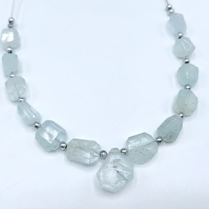 May include: A strand of aquamarine gemstone beads. The beads are a pale blue-green color, with irregular, faceted shapes. The beads are strung on a thin, silver-colored wire, with small silver beads between each gemstone. The background is white.
