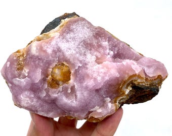 May include: A large, raw, uncut piece of pink rhodochrosite crystal. The stone has a layered appearance with areas of light pink, white, and brown. The rhodochrosite is held in a hand against a white background.