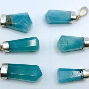 May include: Six turquoise-colored crystal pendants with silver-colored metal caps and loops. The crystals have a faceted, geometric shape. The pendants are displayed on a white surface.
