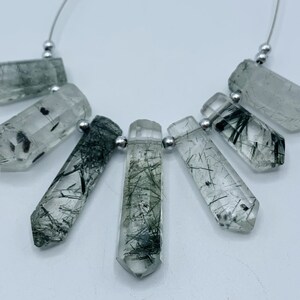 Könnte beinhalten: Eine Sammlung klarer Quarz-Kristall-Anhänger mit schwarzen Turmalin-Einschlüssen. Jeder Kristall hat eine hexagonale Prismenform und ist auf einem Silberdraht mit kleinen Silberperlen aufgefädelt. Die Kristalle variieren in der Größe.