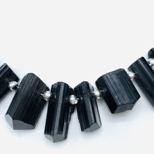 May include: A necklace featuring black tourmaline gemstone beads. The rectangular, polished stones are strung on a thin, silver-colored wire with small silver beads between each stone. The background is plain white.