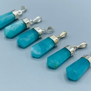 May include: Five blue-green crystal pendants with silver-colored metal caps and loops. The pendants are hexagonal in shape and have a faceted surface. The background is a light blue-gray color.