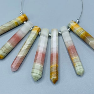 May include: A collection of seven gemstone pendants with a striped pattern in shades of white, pink, yellow, and green. Each pendant is a long, faceted, hexagonal shape, strung on a thin silver wire. The gemstones are displayed against a light gray background.