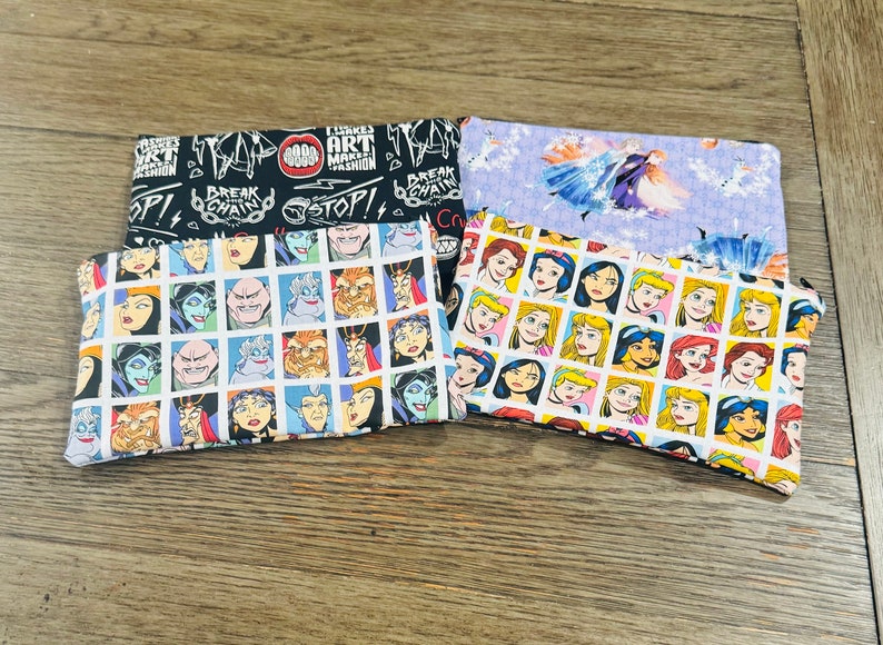 May include: Three fabric pouches with different designs. The top pouch is black with white text that reads "FLIRKS ART MAKES FASHION" and "BREAK THE CHAIN". The middle pouch is light purple with a white silhouette of a princess in a blue dress. The bottom pouch is white with a grid of colorful cartoon images of princesses.