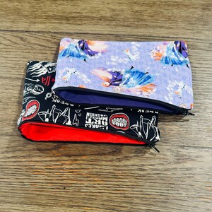 May include: Three zippered pouches with different designs. The top pouch is purple with a white snowflake pattern and a picture of a woman in a blue dress. The middle pouch is black with a white zipper and a red lining. The bottom pouch is black with white text and graphics. The text includes "GOOD", "MAKE", "ART", "FASHION", "LACKED", "BOOM", "GEEKY", "REBEL".