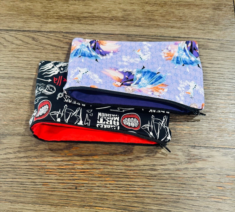 May include: Three zippered pouches with different designs. The top pouch is purple with a white snowflake pattern and a picture of a woman in a blue dress. The middle pouch is black with a white zipper and a red lining. The bottom pouch is black with white text and graphics. The text includes "GOOD", "MAKE", "ART", "FASHION", "LACKED", "BOOM", "GEEKY", "REBEL".