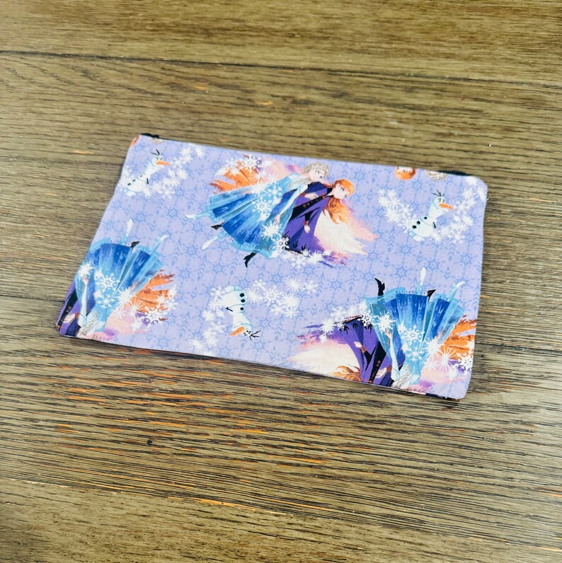 May include: A purple zippered pouch with a repeating pattern of Elsa and Anna from Frozen. The pattern also includes snowflakes and Olaf the snowman.