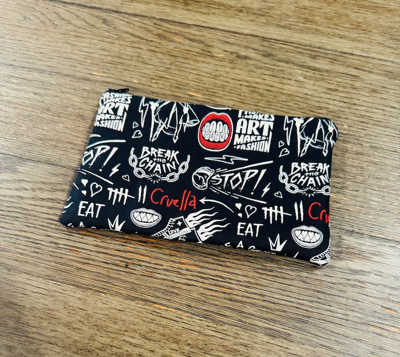 May include: Black zippered pouch with a white and red graphic print. The print features the text "Break the Chain", "Cruella", "Art Makes Fashion", and "Stop!" along with other graphic elements.