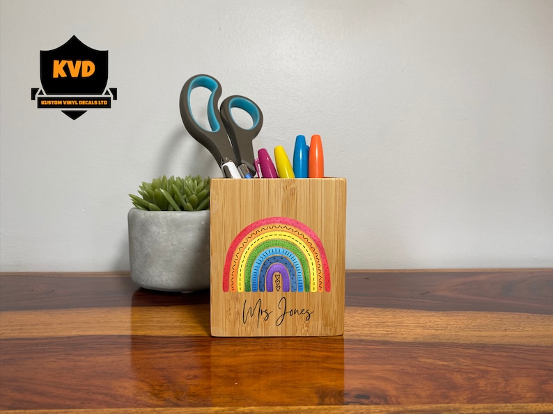Pu&ograve; includere: Un organizer da scrivania in bamb&ugrave; con un colorato design arcobaleno e il nome "Mrs. Jones" sotto. Contiene forbici e pennarelli colorati. A sinistra c'&egrave; una piccola pianta grassa in un vaso grigio.