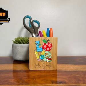 Puede incluir: Un porta lápices de madera con un diseño colorido de "LOVE" que incluye una manzana roja, lápices, un globo terráqueo y libros. El porta lápices tiene el texto "Mrs. Smith" en la parte inferior.