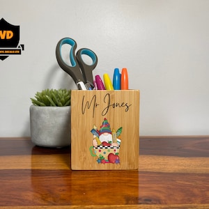 Puede incluir: Un porta lápices de madera con un diseño de gnomo y el nombre "Mr. Jones" impreso en la parte delantera. El porta lápices está lleno de bolígrafos y lápices de colores.