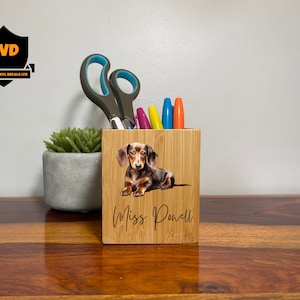 Puede incluir: Un organizador de escritorio de bambú con una imagen impresa de un perro salchicha marrón y negro. El nombre "Miss Powell" está grabado en la parte delantera. El organizador está lleno de bolígrafos y lápices.
