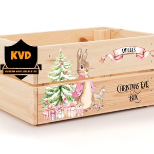 May include: A wooden box with a Christmas Eve Box design. The box is decorated with a pink ribbon, a Christmas tree, a bunny rabbit, and the text "Amelia's Christmas Eve Box".