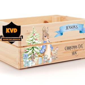 Könnte beinhalten: Eine Holzkiste mit einem Weihnachts-Abend-Box-Design. Die Kiste ist mit einem blauen Band, einem Weihnachtsbaum und einem Hasen verziert. Der Text "Jenson's Christmas Eve Box" ist auf die Kiste gedruckt.
