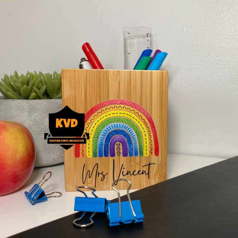 Pu&ograve; includere: Un portapenne in legno con un colorato design arcobaleno e il nome "Mrs Vincent". Il portapenne contiene penne, matite e un righello. Fermagli blu e una mela in primo piano.