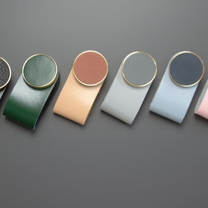 Op de afbeelding: Zeven lederen ladegrepen met messing accenten. De grepen zijn in verschillende kleuren: zwart, groen, beige, grijs, lichtblauw, roze en donkerblauw.