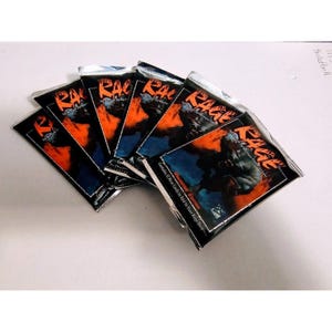 May include: Several packs of "Rage" trading cards are fanned out on a white surface. Each pack features a dark illustration with orange and blue hues, the word "Rage" in bold letters, and text that reads "Contains 12 New Cards to Add to Your Rage Deck."