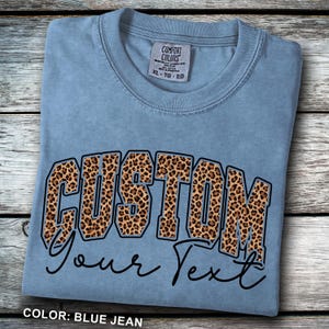 May include: A blue jean Comfort Colors t-shirt with a custom design. The word "CUSTOM" is printed in large, leopard print letters, with the words "Your Text" in a cursive font below. The shirt is laid on a wooden surface.