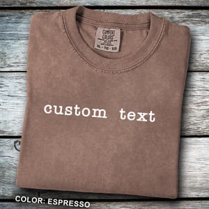 May include: Espresso-colored t-shirt with the words "custom text" in white. The shirt is folded neatly, with a tag visible at the neckline. The shirt is made of a soft material. The color is espresso.