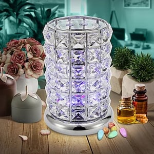 May include: A crystal lamp with a silver base and a cylindrical design. The lamp emits a soft purple glow, and is surrounded by candles, essential oil bottles, and decorative stones. The lamp is placed on a wooden surface.