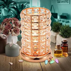 May include: A rose gold-coloured, cylindrical candle holder with a base, decorated with sparkling, clear crystals. The candle holder is surrounded by decorative items, including small glass bottles, candles, and colourful stones, creating a warm ambience.