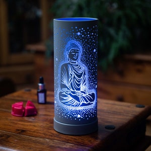 May include: A white ceramic oil diffuser with a blue light and a design of a meditating Buddha. The diffuser has a star-like pattern and a round base.
