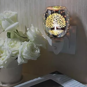 May include: A glass plug-in essential oil diffuser featuring a tree of life design. The diffuser is illuminated, giving off a warm light. White roses in a white vase are in the background. The diffuser is plugged into a white wall socket.