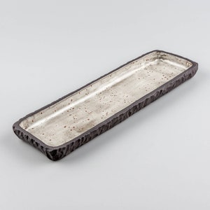 Op de afbeelding: Een lange, rechthoekige serveerschaal van keramiek met een getextureerde, donkerbruine rand en een gespikkelde, lichtgrijze binnenkant.