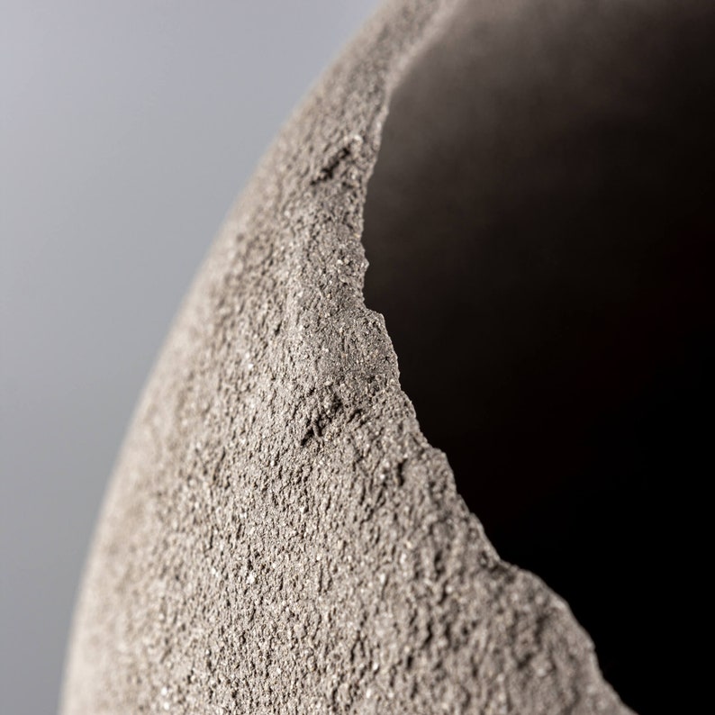 Pode incluir: Close-up de um vaso de cer&acirc;mica cinza texturizado com uma abertura &aacute;spera e irregular. O vaso tem um aspecto r&uacute;stico e feito &agrave; m&atilde;o.