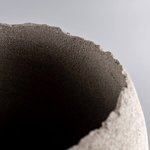 Pode incluir: Um close-up do topo de um vaso de cer&acirc;mica cinza com uma superf&iacute;cie &aacute;spera e texturizada. O vaso tem uma abertura redonda e larga.
