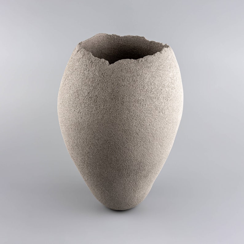 Pode incluir: Um vaso de cer&acirc;mica cinza com uma superf&iacute;cie &aacute;spera e texturizada e uma abertura irregular e desigual na parte superior.