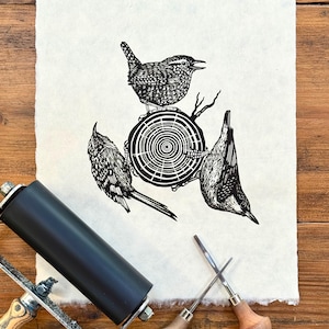 May include: A black and white linocut print of three birds perched on a tree stump. The print is on textured, off-white paper. A black ink roller and two carving tools are in the foreground, suggesting the printmaking process.
