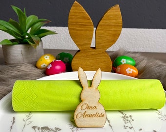 May include: Wooden Easter bunny napkin holder with the name "Oma Annelise" engraved on it. A green napkin is placed on a white plate with a floral design. Decorative Easter eggs and a wooden bunny silhouette are in the background.