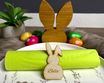 May include: A wooden bunny napkin ring with the name "Bella" engraved on it. The ring is placed on a green napkin on a white plate with a floral design. There are Easter eggs in the background.