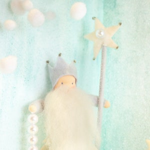 Puede incluir: Una muñeca de fieltro blanco con una corona gris, barba blanca y alas blancas. La muñeca sostiene una varita blanca en forma de estrella con una perla en el centro. La muñeca está de pie sobre un fondo azul claro.