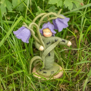 Könnte beinhalten: Eine Filzpuppe mit grünem Kleid und einer Blumenkrone steht in einem Bett aus grünem Gras mit violetten Blumen. Die Puppe hat ein hellbraunes Gesicht und blonde Haare. Auf dem Kleid der Puppe und dem Sockel der Puppe befinden sich kleine schwarze Filz-Ameisen.