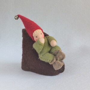 May include: A small felt gnome doll wearing a red hat with a bell sits on a brown felt chair. The gnome is wearing a green felt outfit and has a white felt face.