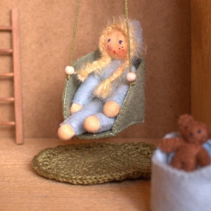 May include: A doll with blonde hair sits on a green swing in a dollhouse. The doll is wearing a blue outfit and has a wooden face. There is a wooden ladder and a brown rope swing in the background.