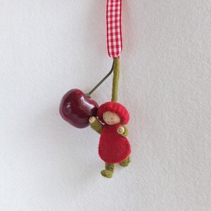 May include: A felt doll with a red dress and a red hat holds a red cherry. The doll is hanging from a green stem with a red and white checkered ribbon.
