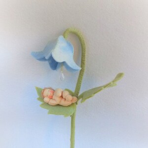 以下が含まれることがあります： 手作りのフェルトフラワー。薄い青色のベル型の花と緑色の茎があります。小さなベージュ色の赤ちゃん人形が緑の葉の上に寄り添っています。透明なクリスタルが花からぶら下がっています。