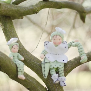 Puede incluir: Tres mu&ntilde;ecas de fieltro hechas a mano vestidas con atuendos verdes y blancos est&aacute;n posadas en ramas de &aacute;rboles. Las mu&ntilde;ecas tienen caras pintadas y llevan sombreros.