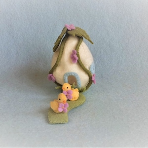 Op de afbeelding: Een wit vilten eivormig huis met een groen vilten dak en paarse bloemen. Twee gele vilten eendjes staan voor het huis op een groen vilten pad.