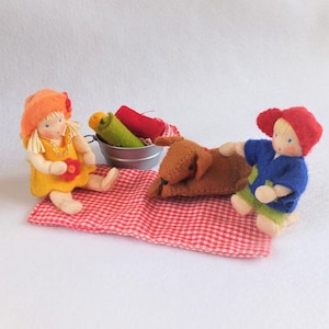 Op de afbeelding: Twee poppen van vilt, een met een rode hoed en een blauw shirt, de andere met een oranje hoed en een gele jurk, zitten op een rood-wit geblokt kleed met een bruine vilt hond die tussen hen ligt. Een kleine metalen emmer met groene en rode vilt items staat op de achtergrond.